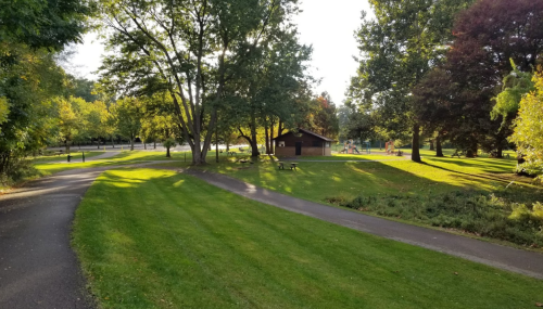A peaceful park scene with green grass, trees, and a wooden shelter, featuring winding paths and a playground in the background.