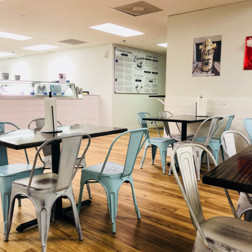 A bright, modern café with wooden floors, metal chairs, and a menu board on the wall. Tables are set for customers.