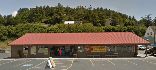 A single-story building with a red roof, featuring a sign for a museum, surrounded by trees and parked cars.