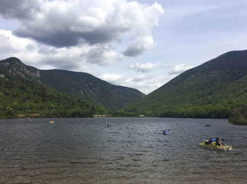 A serene lake surrounded by mountains, with people kayaking and cloudy skies above.