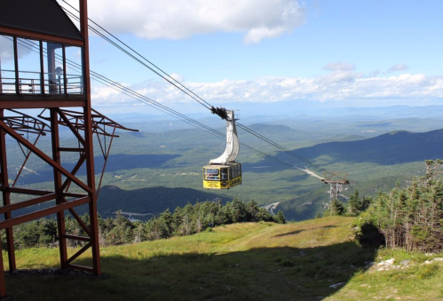A cable car glides over a lush green landscape, with mountains and a blue sky in the background.