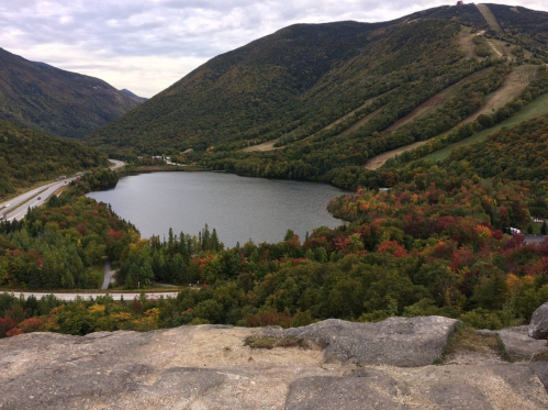 A scenic view of a lake surrounded by colorful autumn foliage and mountains under a cloudy sky.