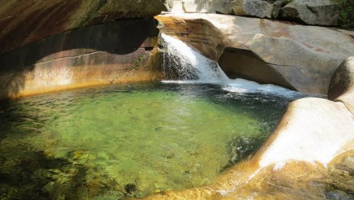 A serene natural pool with clear green water, surrounded by smooth rocks and a gentle waterfall.