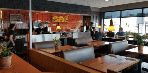 A cozy restaurant interior with booths, plants, and patrons dining, while staff prepare food in the background.