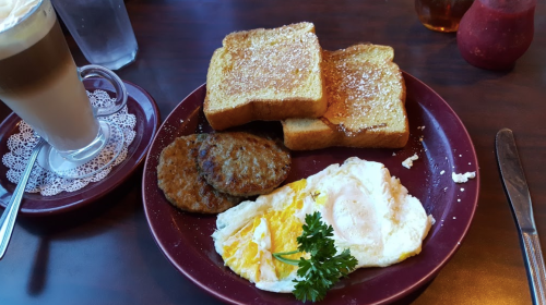 A plate with two slices of French toast, a fried egg, sausage patties, and a cup of coffee on the side.