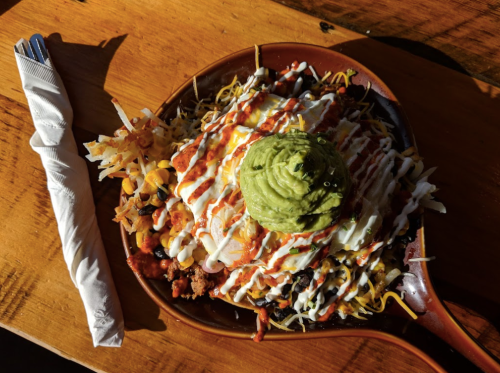 A sizzling platter of nachos topped with cheese, guacamole, sour cream, and drizzled with sauce, served on a wooden table.
