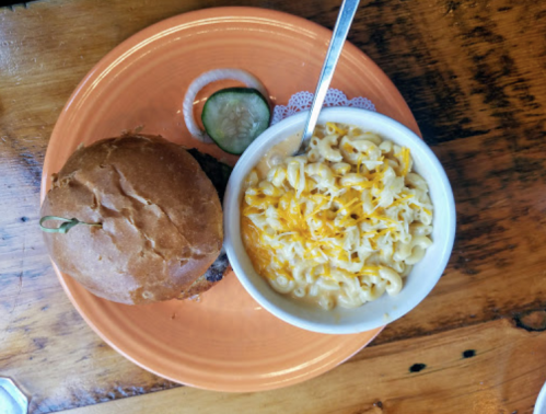 A plate with a burger, a pickle slice, and a bowl of macaroni and cheese topped with shredded cheese.