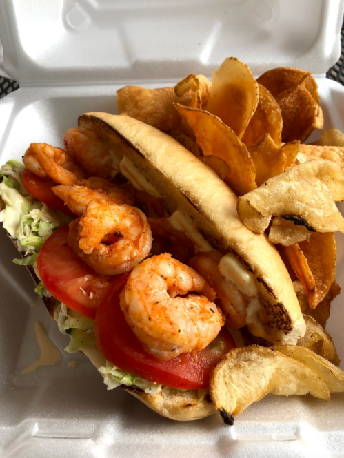 A shrimp sandwich with lettuce, tomato, and sauce, served with crispy potato chips on the side.