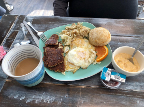 A plate with two fried eggs, hash browns, a biscuit, orange slice, and a bowl of grits, alongside a cup of coffee.