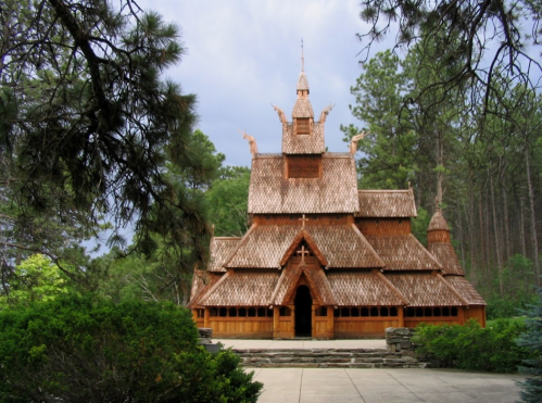 A wooden church with intricate architecture, surrounded by trees and greenery under a cloudy sky.