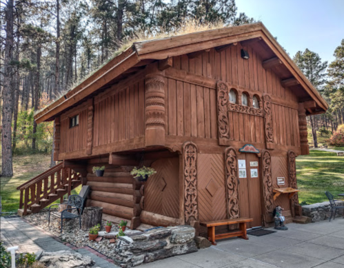 A rustic wooden building with intricate carvings, a thatched roof, and surrounded by trees and greenery.