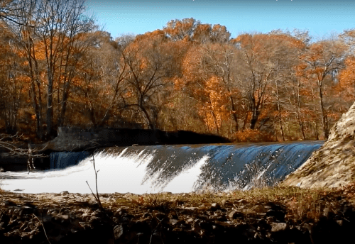 A serene waterfall cascades over rocks, surrounded by vibrant autumn foliage and bare trees.