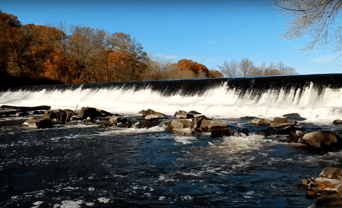 A waterfall cascades over a dam, surrounded by rocks and autumn trees under a clear blue sky.