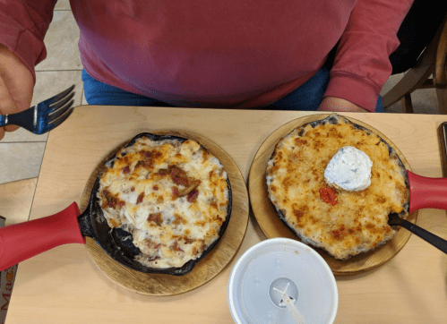 Two sizzling skillets of cheesy dishes on wooden plates, one topped with a dollop of sour cream and the other with a red sauce.