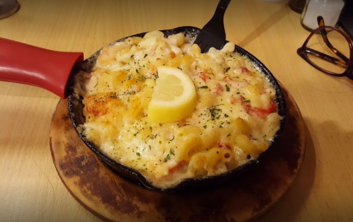 A skillet of baked macaroni and cheese topped with a lemon slice and herbs, served on a wooden board.