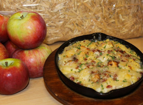 A cast-iron skillet filled with creamy macaroni and cheese, topped with herbs and bacon, beside fresh red apples.