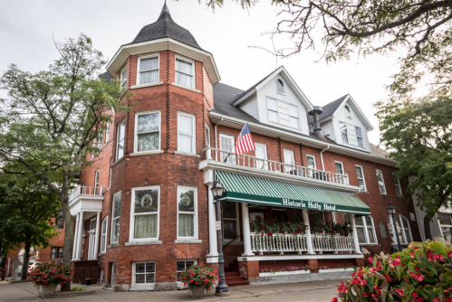 Historic brick hotel with a tower, green awning, and American flag, surrounded by trees and flower planters.