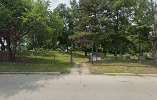 A cemetery entrance with a sign, surrounded by trees and gravestones on a grassy hill.