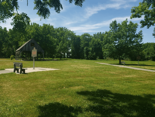 A grassy park with a wooden shelter, a bench, and trees under a clear blue sky.