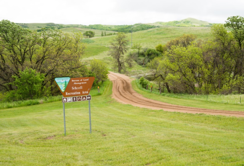Sign for Shell Recreation Area at a dirt road entrance, surrounded by green hills and trees.