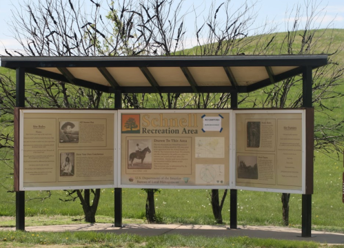 Information sign for Schnell Recreation Area, featuring historical photos and details about the area. Green landscape in background.