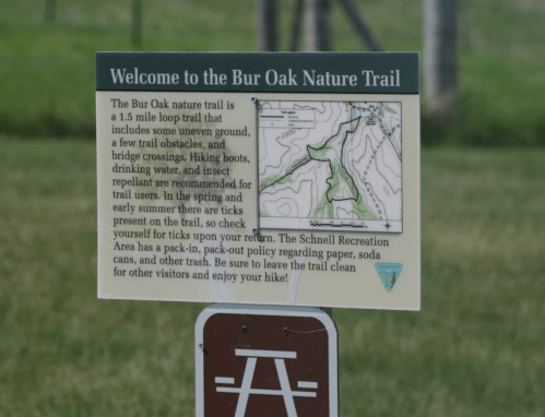Sign for Bur Oak Nature Trail detailing trail features, safety tips, and a map, set against a grassy background.