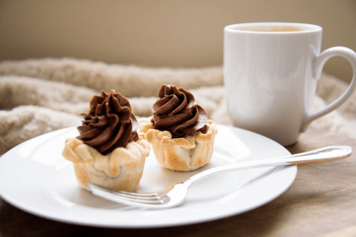 Two chocolate-filled pastry cups on a white plate, accompanied by a cup of coffee. Soft, cozy background.