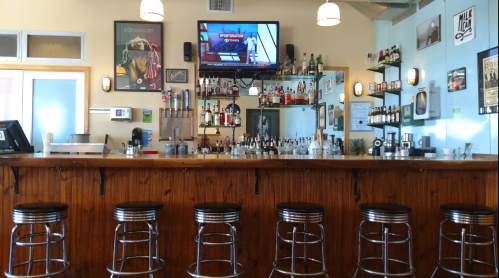 A bar with a wooden counter, empty stools, shelves of liquor, and a TV displaying a show in a bright, inviting space.