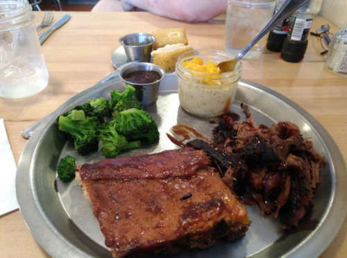 A metal plate with barbecue meat, broccoli, cornbread, and sides of sauce and macaroni in a jar.