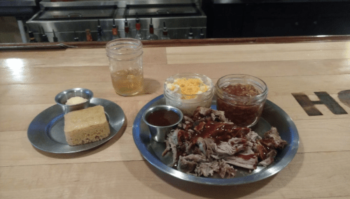 A plate with pulled pork, barbecue sauce, coleslaw, baked beans, cornbread, and a drink on a wooden table.