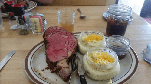 A plate with a large slice of prime rib, two jars of mashed potatoes topped with cheese, and drinks on a wooden table.