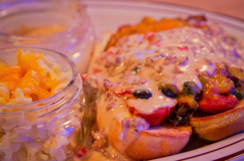 A plate featuring a cheesy sandwich topped with vegetables, alongside two jars of macaroni and cheese and coleslaw.