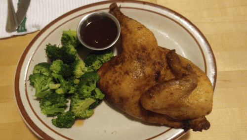 A plate with a roasted chicken half, served with steamed broccoli and a small cup of barbecue sauce.