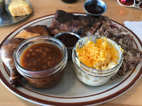 A plate of barbecue featuring chicken, ribs, pulled pork, baked beans, and macaroni and cheese, with sauce on the side.