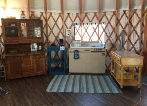 Interior of a yurt featuring a vintage kitchen setup with cabinets, a stove, and a striped rug on wooden flooring.