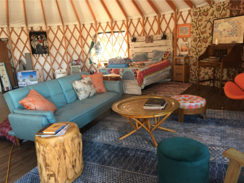Cozy yurt interior with a blue sofa, round coffee table, colorful decor, a bed, and a piano. Warm, inviting atmosphere.