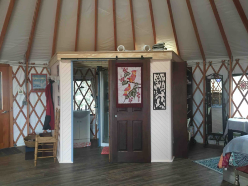 Interior of a yurt featuring wooden beams, a bathroom area, and decorative elements like a bird-themed wall hanging.