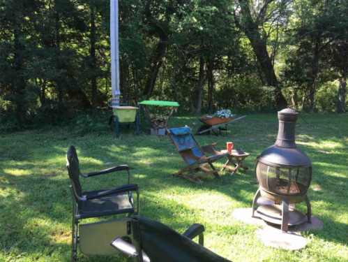 A peaceful outdoor scene with chairs, a fire pit, and a wheelbarrow in a green, wooded area.