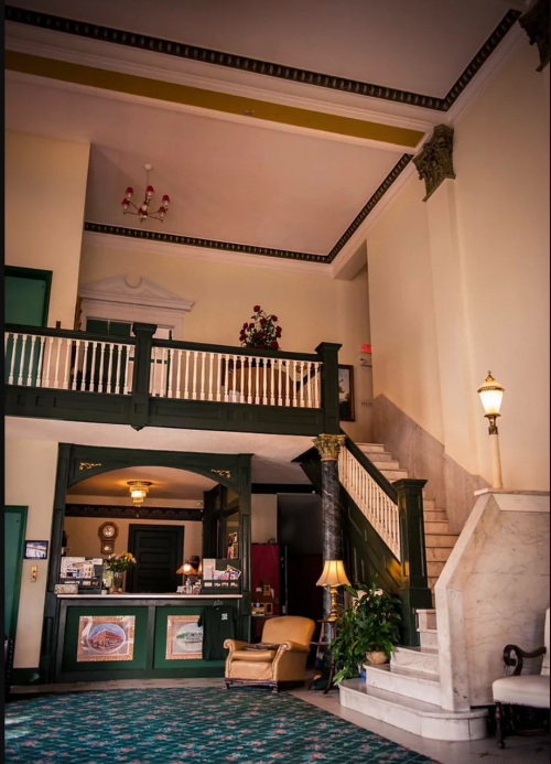 Interior of a vintage hotel lobby featuring a staircase, reception desk, and elegant decor with green and gold accents.