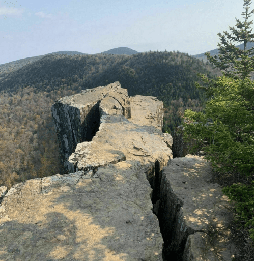 A rocky cliff edge overlooking a forested mountain landscape under a clear sky.