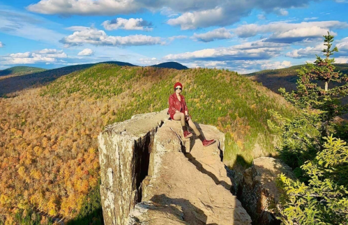 A person sits on a rocky ledge overlooking a colorful autumn landscape with rolling hills and a blue sky.