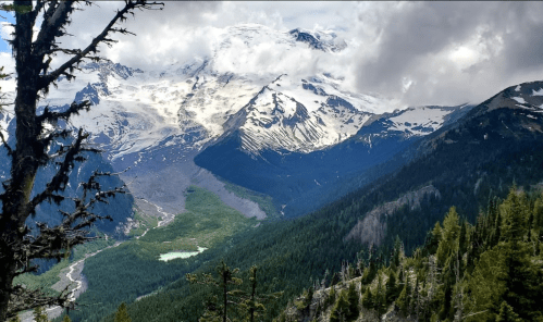 A scenic mountain landscape with snow-capped peaks, lush green valleys, and a cloudy sky.
