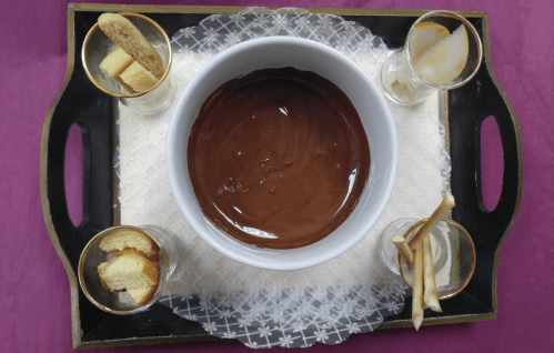 A tray with a bowl of melted chocolate surrounded by glasses of fruit and breadsticks for dipping.