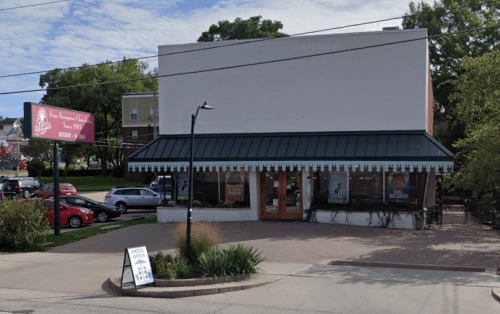 A storefront with a green awning, large windows, and a sign indicating it's open, surrounded by parked cars and greenery.
