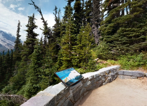 A scenic viewpoint with a stone wall, surrounded by tall trees and mountains, featuring an informational sign.