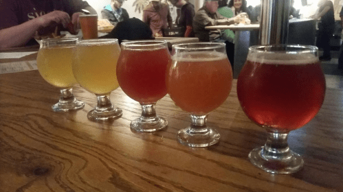 Five glasses of colorful craft beer lined up on a wooden table, with people socializing in the background.