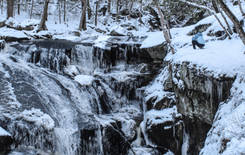 A snowy waterfall cascades over rocks, with a person crouched nearby, surrounded by trees in a winter landscape.