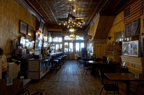 Interior of a vintage bar with wooden floors, a long counter, and decorative lighting, featuring an American flag on the wall.