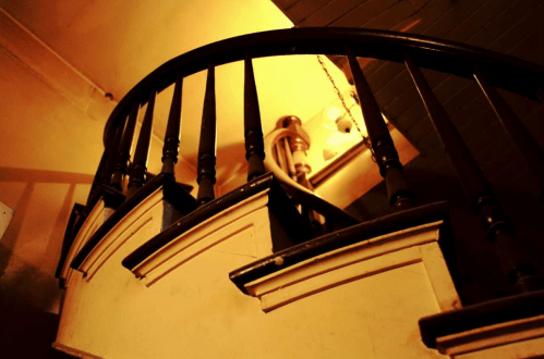 A spiral staircase with wooden railings, illuminated by warm light from a hanging lamp above.