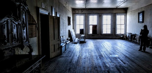 A dimly lit, empty room with wooden floors, large windows, and scattered furniture against the walls.